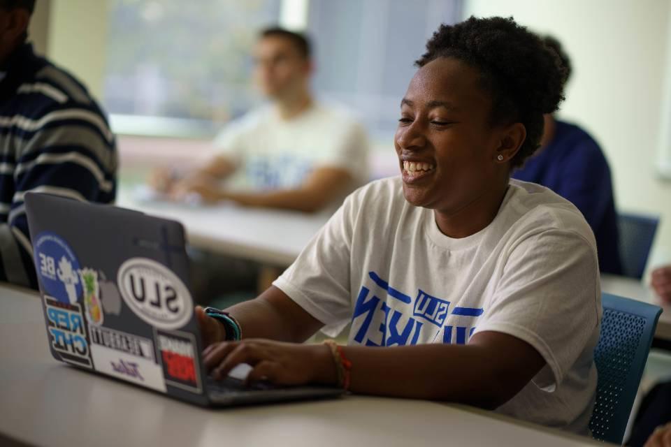 A student wearing a Billiken t-shirt sits in a classroom, smiling while typing on a laptop.